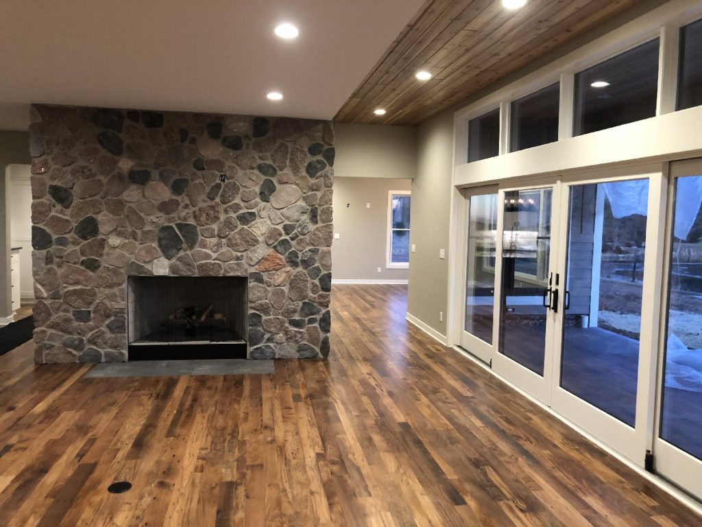 Image of family room, in custom built home, featuring floor to ceiling stone fireplace and wood flooring and large windows.